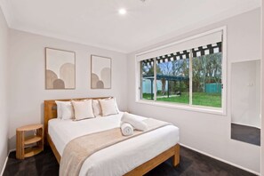 A bedroom with a bed, a small wooden side table, two framed wall art pieces, a large window with a view of the outdoors, and a door. - Casa Cypress, Home Near Bowral with Garden and BBQ (Bowral)