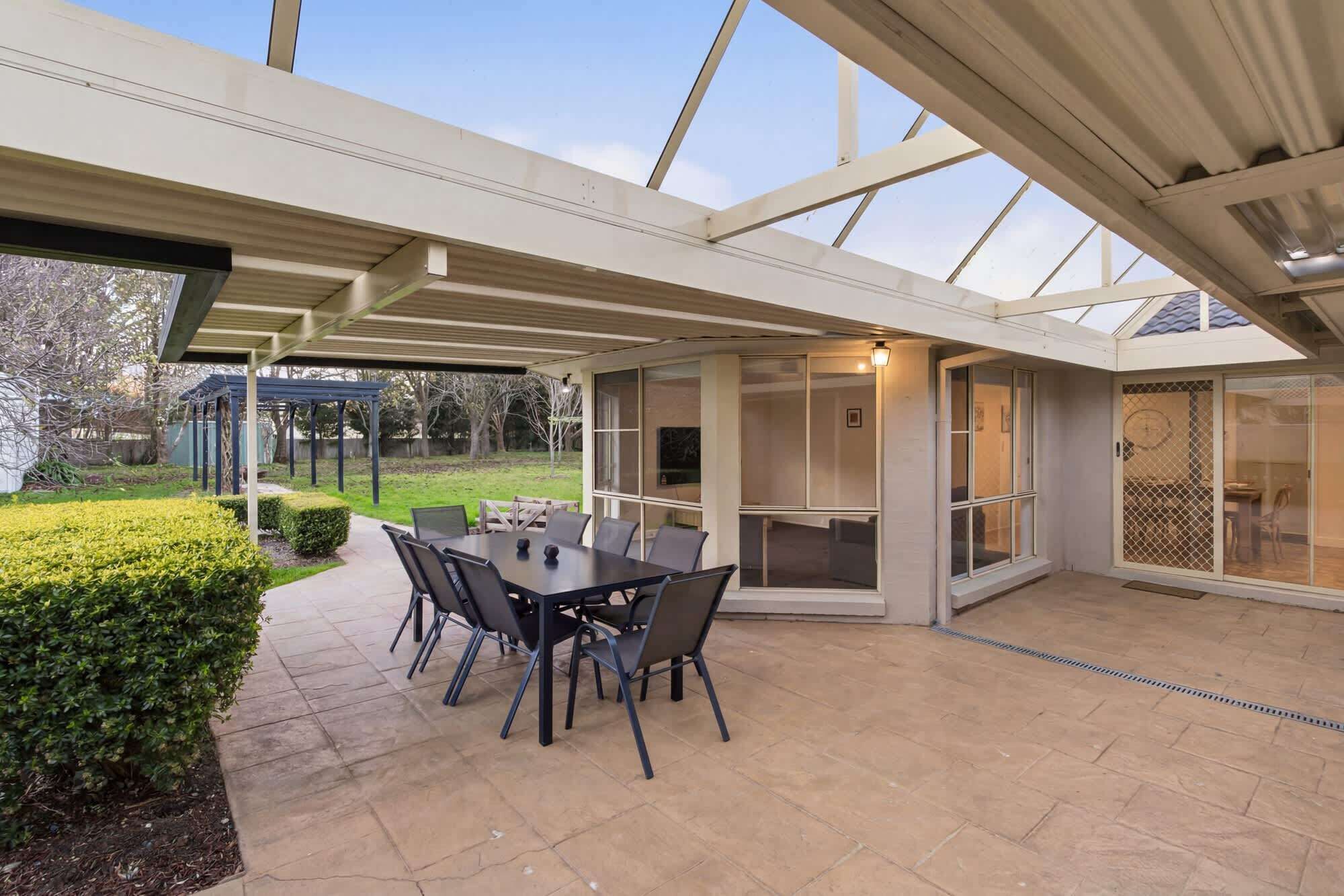 An outdoor patio with a table and chairs, a covered area with a glass enclosure, and a view of the garden.