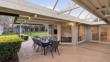 An outdoor patio with a table and chairs, a covered area with a glass enclosure, and a view of the garden.
