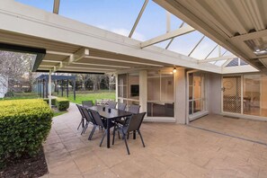 An outdoor patio with a table and chairs, a covered area with a glass enclosure, and a view of the garden. - Casa Cypress, Home Near Bowral with Garden and BBQ (Bowral)