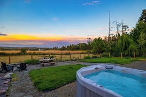 A hot tub in a backyard with a view of the sunset, a picnic table, and chairs.