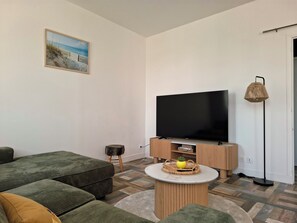 Una sala de estar con televisor de pantalla plana, sofás verdes, un taburete pequeño, una mesa de centro redonda y una lámpara de pie.