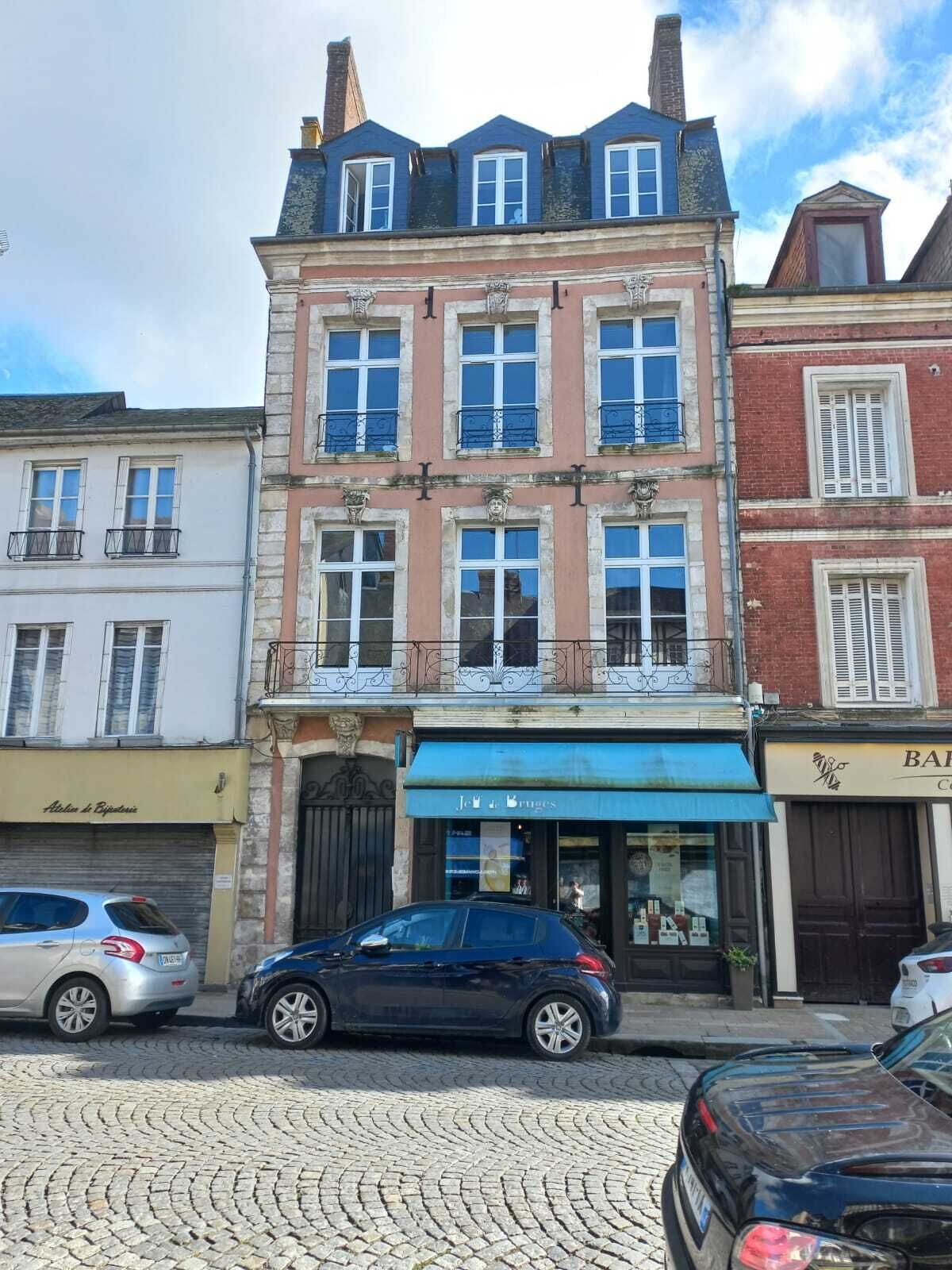A multi-story building with a row of windows on the ground floor, a storefront with a blue awning, and parked cars on a cobblestone street.