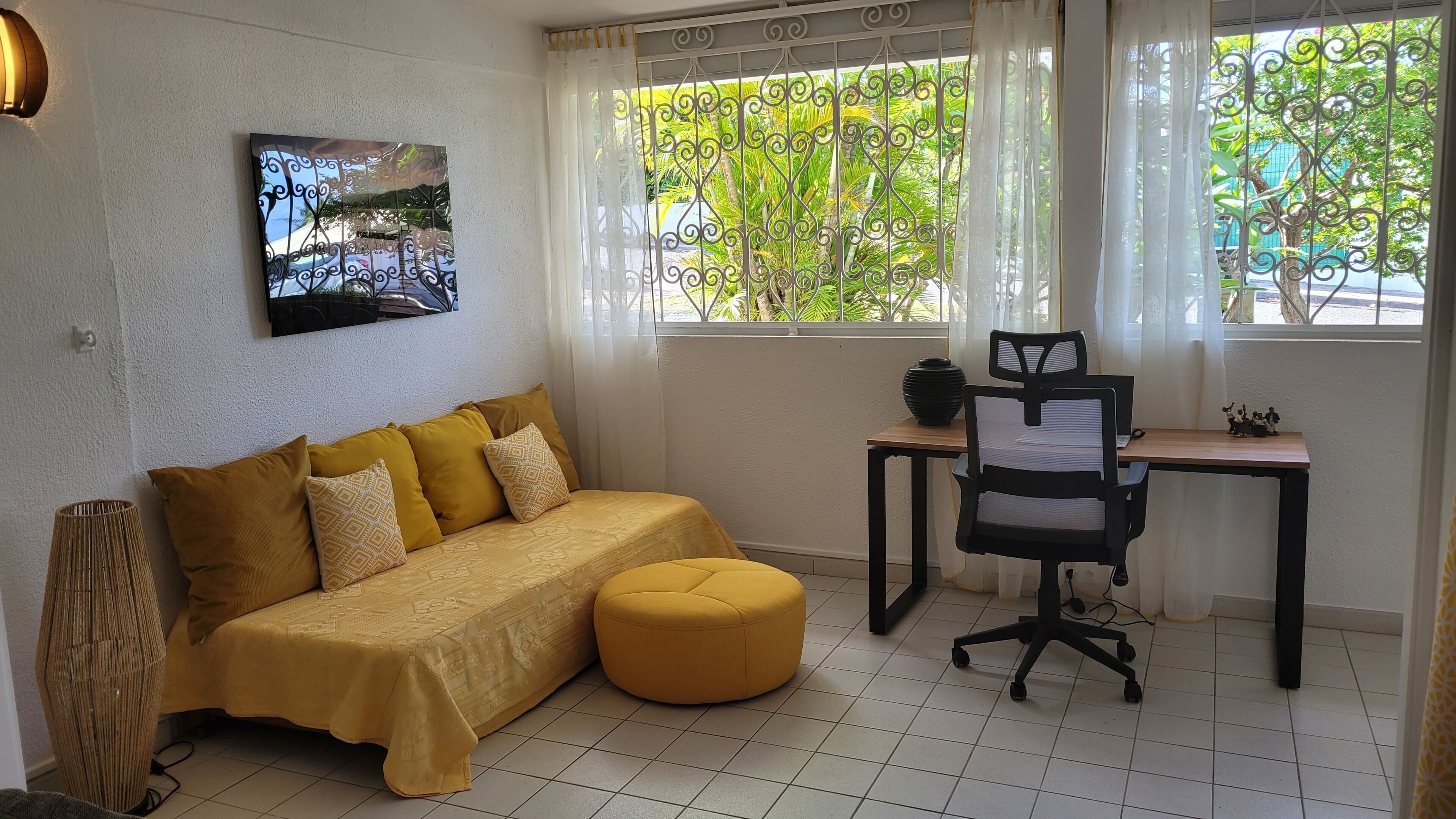 A room with a sofa, a small round ottoman, a desk with a chair, a wall-mounted picture, and a window with a view of greenery.