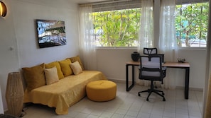 Una habitación con sofá, un otomanillo redondo, un escritorio con silla, un cuadro en la pared y una ventana con vistas a zonas verdes.