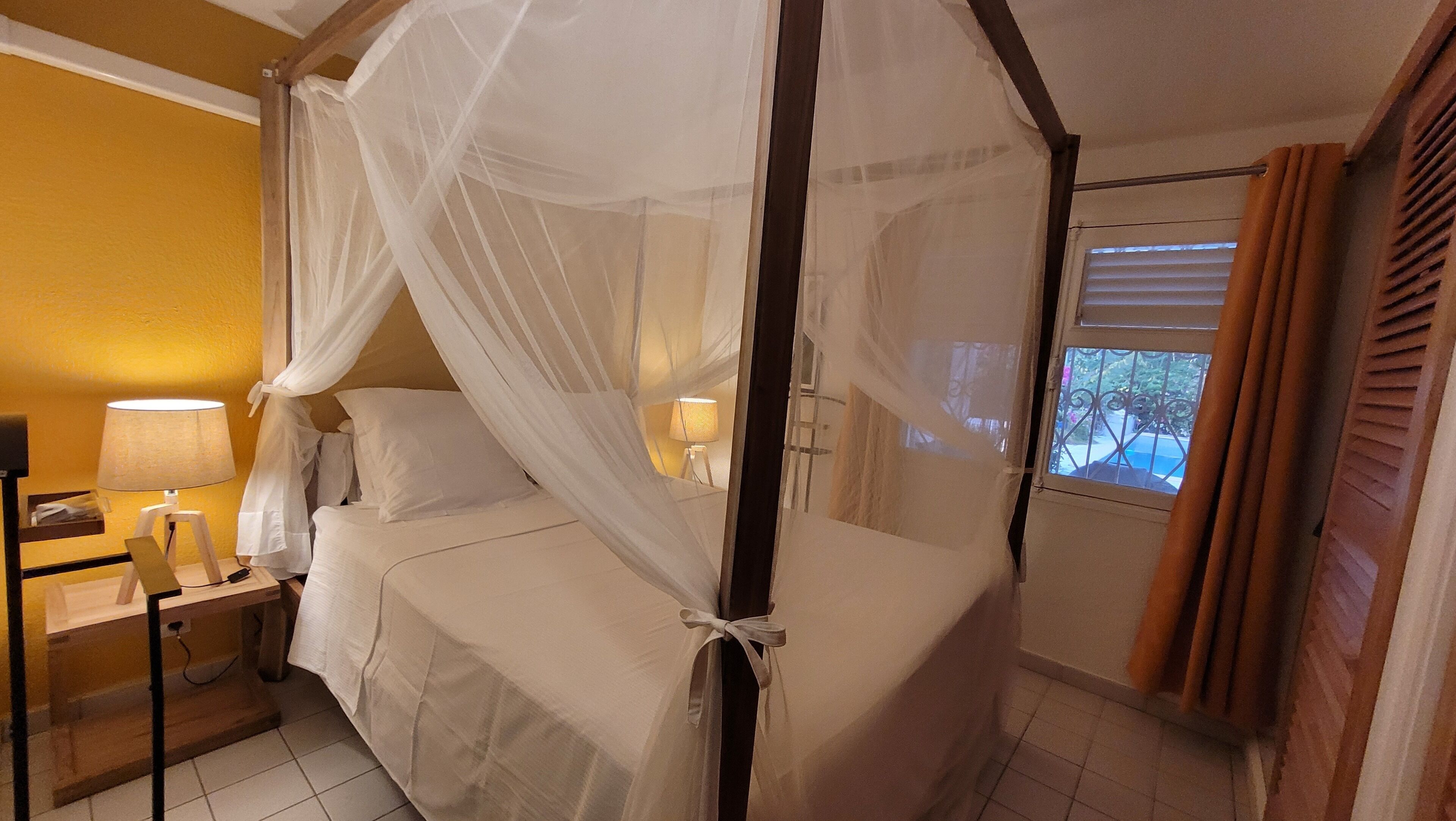 Ein Zimmer mit einem Bett unter einem Baldachin, einem Nachttisch mit Lampe und einem Fenster mit Vorhängen.