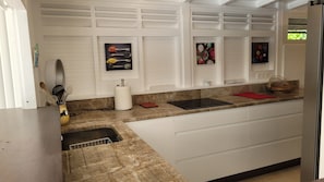 A kitchen with white cabinets, a granite countertop, a black induction cooktop, a red cutting board, and a small window with a view of greenery.