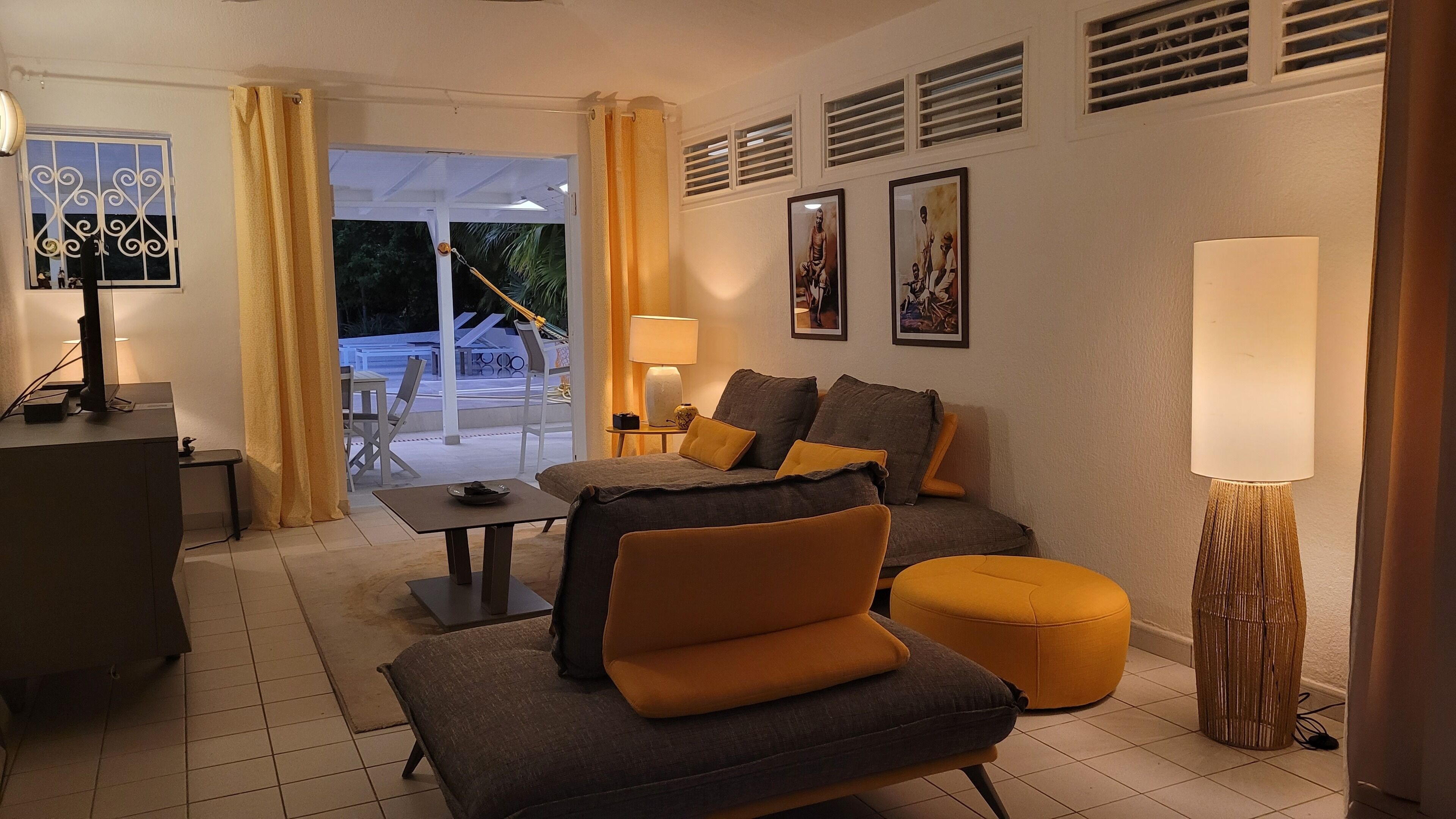 Una sala de estar moderna con sofá, reposapiés y una mesa con lámpara. Hay una ventana con cortinas, un cuadro en la pared y vistas al exterior.