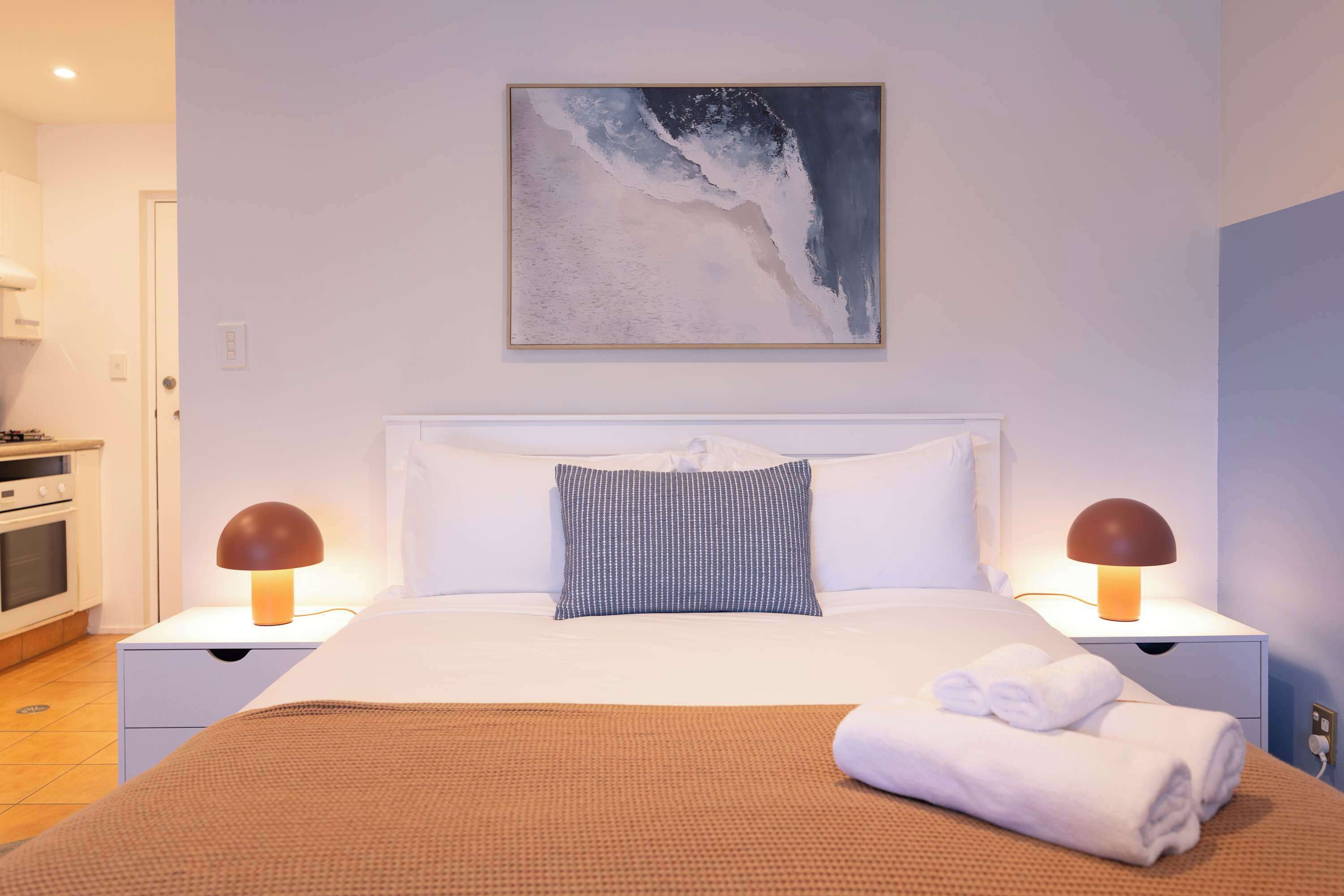 A hotel room with a bed, two bedside lamps, a painting, and a neatly made bed with towels.