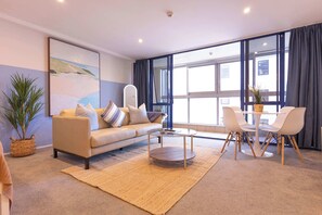 Ein modernes Wohnzimmer mit Sofa, Couchtisch und Sesseln, einem großen Fenster mit Vorhängen und einem Gemälde an der Wand.
