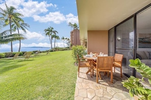 Une terrasse avec une table et des chaises, une pelouse avec des fauteuils de jardin et une vue sur l’océan, bordée de palmiers.
