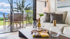 A living room with a glass door leading to an outdoor area, a wooden table with a wine bottle, glasses, and a book, and a sofa with cushions. - Ocean-view 1BR condo with pool, AC and beach access-Maalaea Banyans 112 (Wailuku)