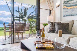 Ein Wohnzimmer mit Glastür zum Außenbereich, ein Holztisch mit Weinflasche, Gläsern und einem Buch sowie eine Couch mit Kissen.