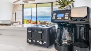 A kitchen with a toaster, a coffee maker, and a view of the ocean through a large window. - Ocean-view 1BR condo with pool, AC and beach access-Maalaea Banyans 112 (Wailuku)