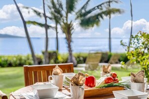 A table set outdoors with a laptop, cups, and a wooden board with vegetables. - Ocean-view 1BR condo with pool, AC and beach access-Maalaea Banyans 112 (Wailuku)