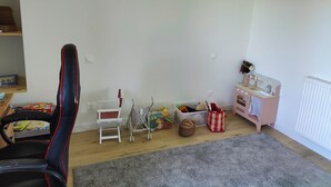 Ein Kinderzimmer mit einem Gaming-Sessel, einem kleinen weißen Stuhl, einer Spielküche und einem grauen Teppich.