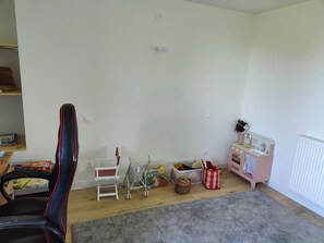 Ett lekrum för barn med en gamingstol, en liten vit stol, ett leksakskök och en grå matta.