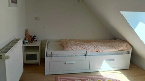 Ein kleines Schlafzimmer im Loft-Stil mit schrägen Wänden, einem Einzelbett mit Blumenmuster, einem Heizkörper, einem Regal mit Stofftieren und einem gerahmten Bild an der Wand.