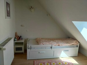 Ett litet, vitt sovrum i loftstil med ett sluttande tak, en enkel säng med ett blommigt överkast, ett element, en hylla med gosedjur och en inramad tavla på väggen.