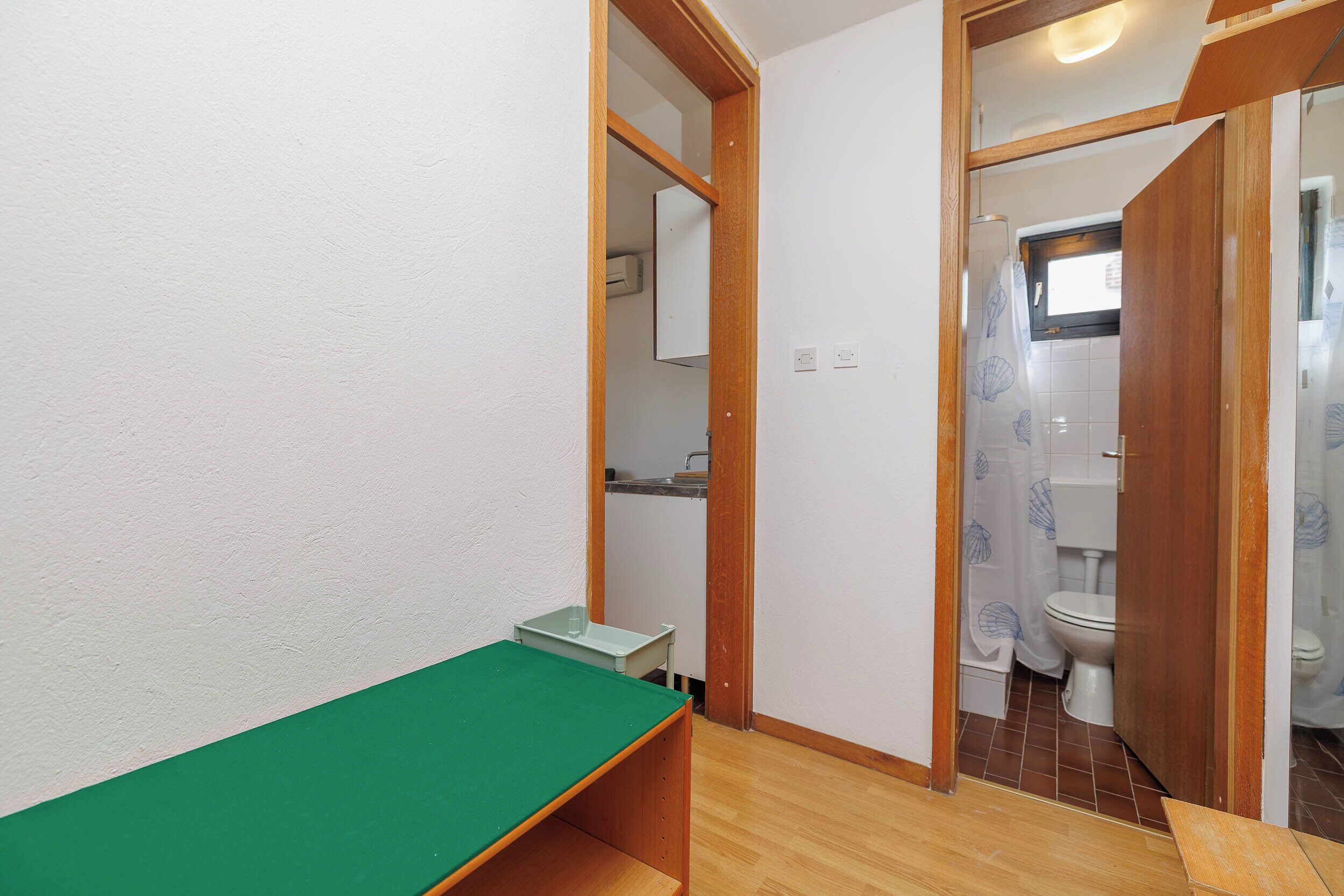Una habitación pequeña con una mesa verde, un banco de madera, una pared blanca y una puerta que da a un baño con inodoro y cortina de ducha.