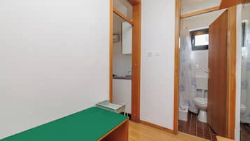 Una habitación pequeña con una mesa verde, un banco de madera, una pared blanca y una puerta que da a un baño con inodoro y cortina de ducha.