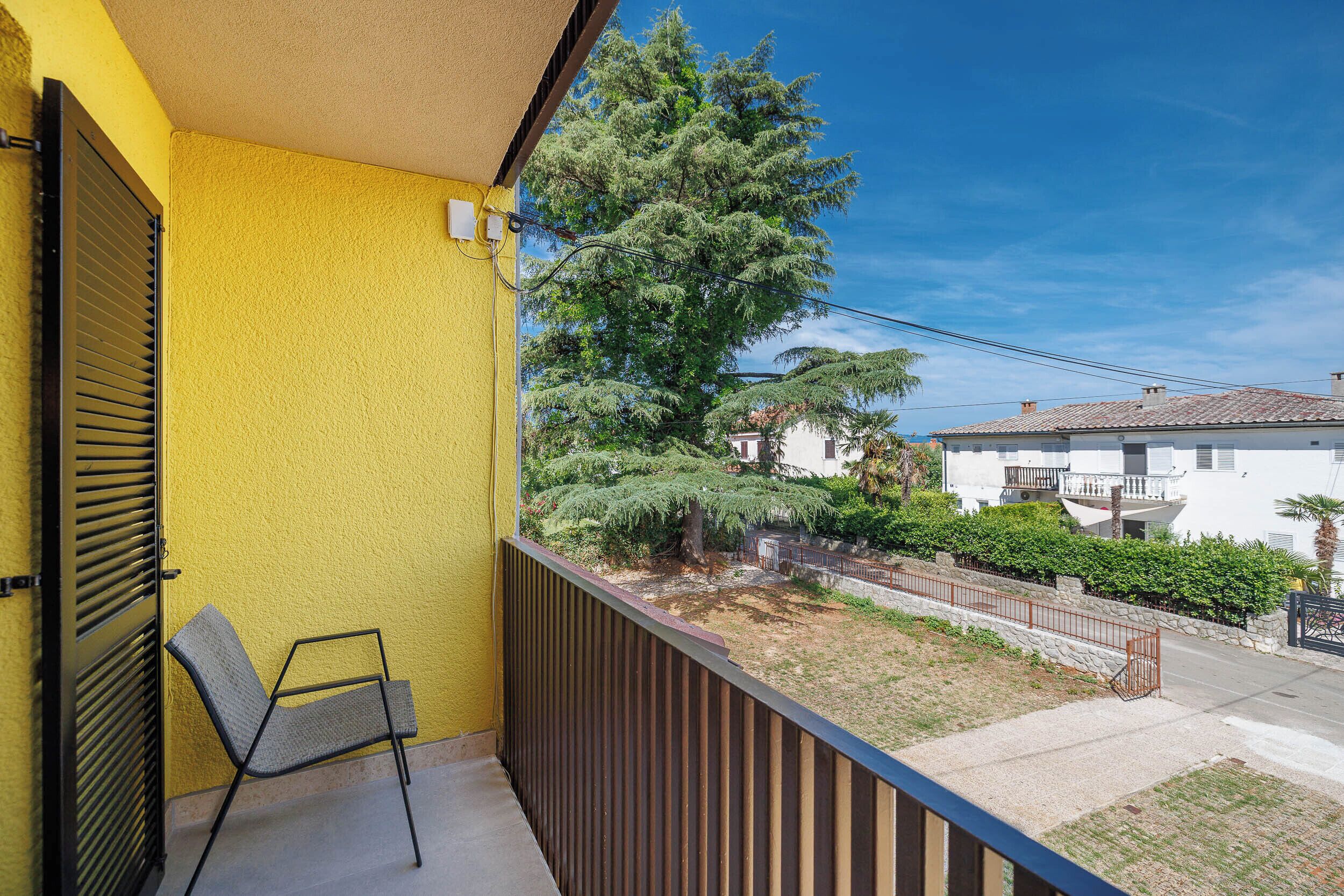 Un balcón con una silla, una pared amarilla y vista a una zona residencial con árboles y casas.