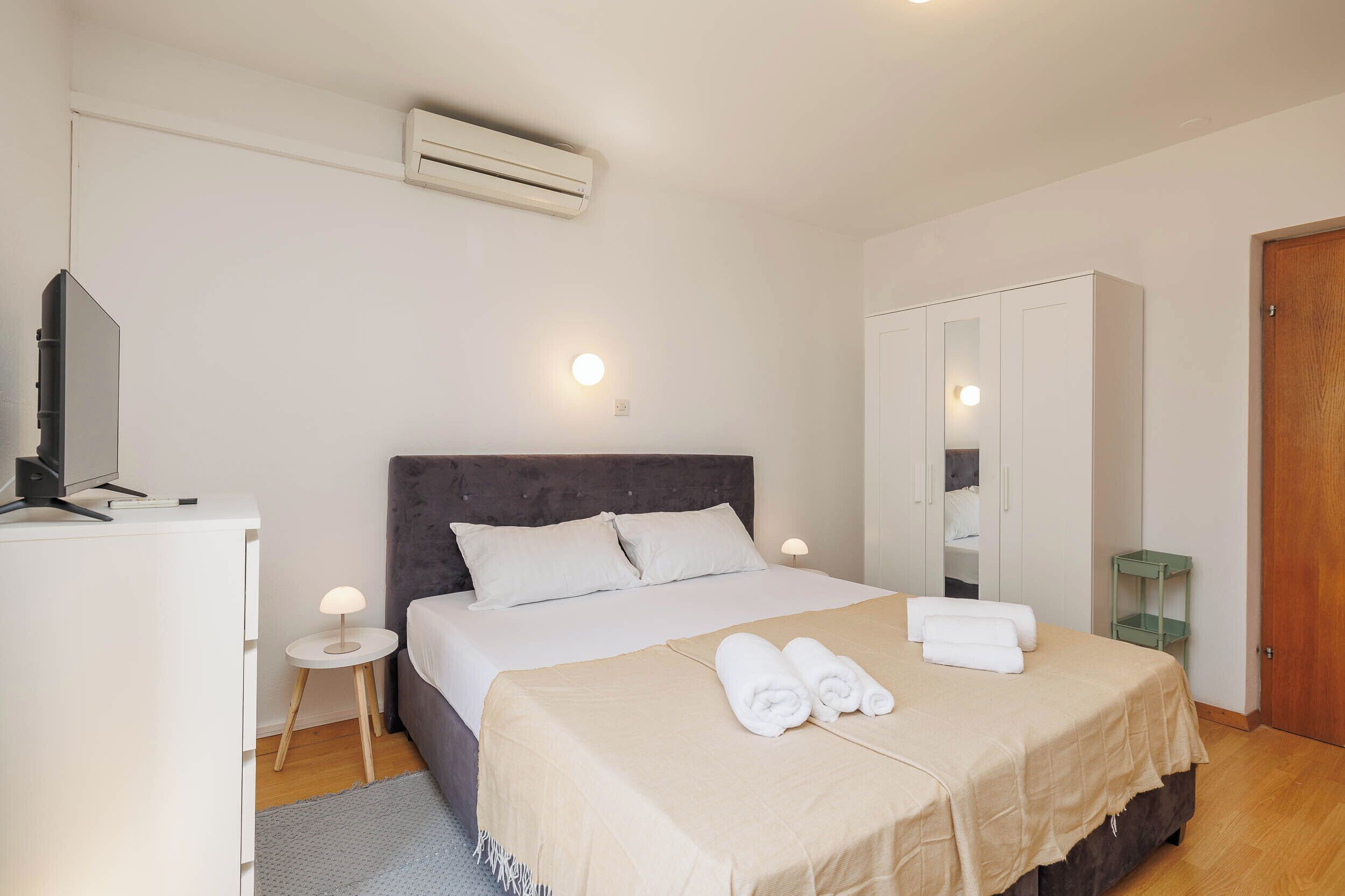 A hotel room with a bed, bedside tables, a wardrobe, an air conditioning unit, and a television.