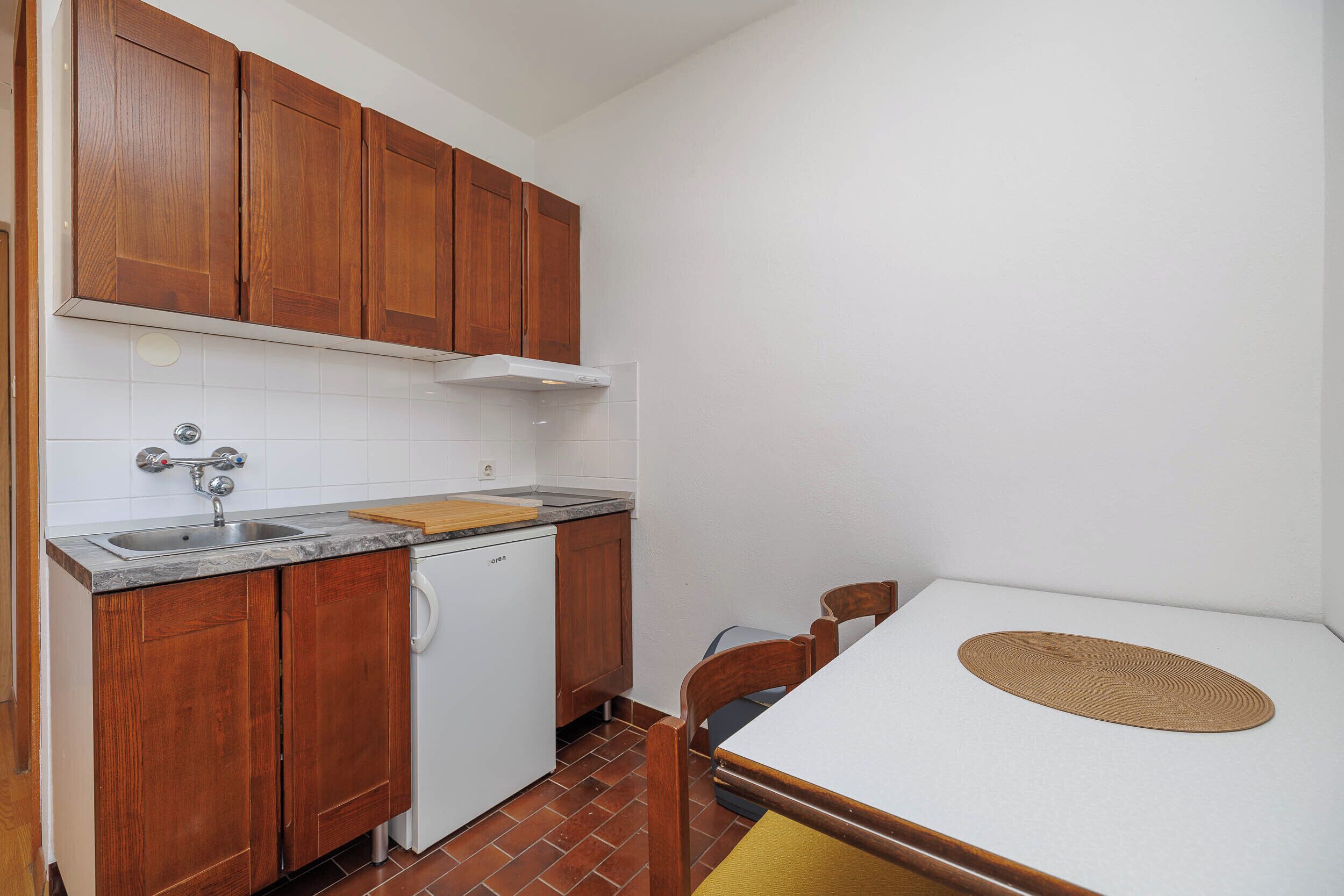 Cocina compacta con armarios de madera, encimera blanca, un lavamanos y una pequeña mesa con un mantel tejido.