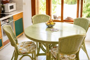 A round dining table with chairs, a bowl of fruit, and a kitchen area with a microwave and cabinets. - Villa Günbatımı (Kaş)