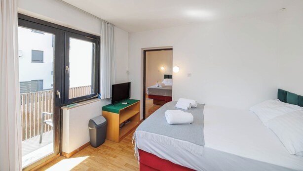 A hotel room with a bed, a TV on a stand, a small desk, and a balcony with a view. - Charming Studio Rental in Malinska with Garden View (Malinska-Dubašnica)