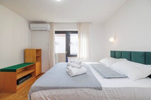 A hotel room with a bed, a wooden shelf, a window with curtains, and an air conditioning unit. - Charming Studio for 4 persons in Malinska (Malinska-Dubašnica)