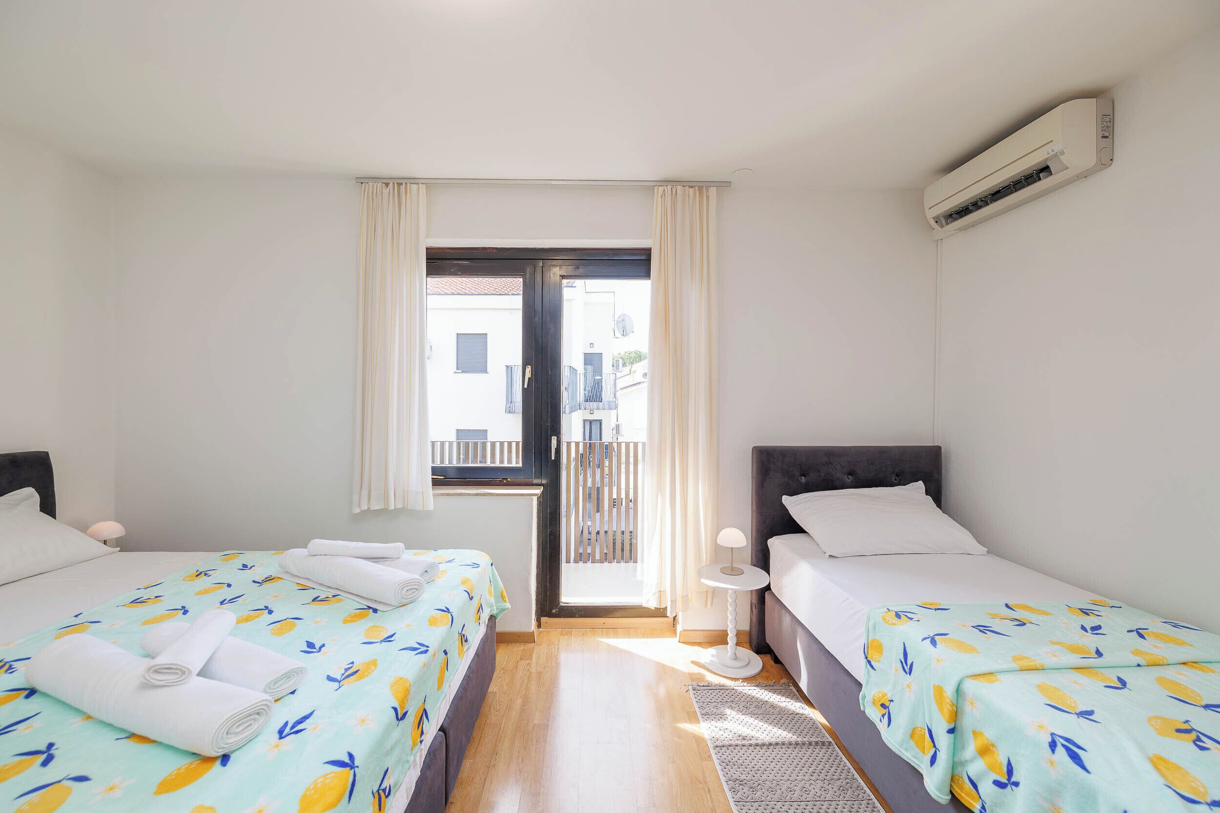 Une chambre avec deux lits, une fenêtre avec des rideaux, une petite table avec une lampe et un appareil de climatisation.