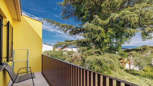 Un balcón con una pared amarilla, una silla de metal y vistas a árboles y colinas.