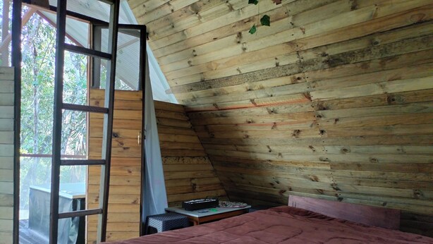 A bedroom with a wooden sloped ceiling, a bed with a red quilt, a small table, and a window with a view of trees. - cabaña con baño interno, en el balcón jacuzzi (Quebradanegra)