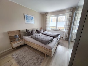 A bedroom with a bed, bedside tables, a lamp, a window with curtains, and a painting on the wall. - 5 - Schröder Apartment - Comfortable vacation rental in Zingst with a terrace and garden (Zingst)