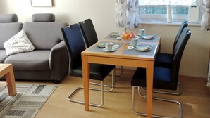 A dining area with a table set for two, a grey sofa, and a window with curtains. - 5 - Schröder Apartment - Comfortable vacation rental in Zingst with a terrace and garden (Zingst)