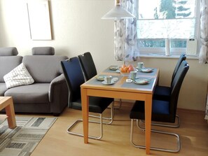 Ein Essbereich mit Tisch für zwei Personen, einem grauen Sofa und einem Fenster mit Vorhängen.