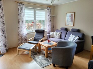Ein Wohnzimmer mit einer Couch, zwei Sesseln, einem Couchtisch und einem Fenster mit Vorhängen.