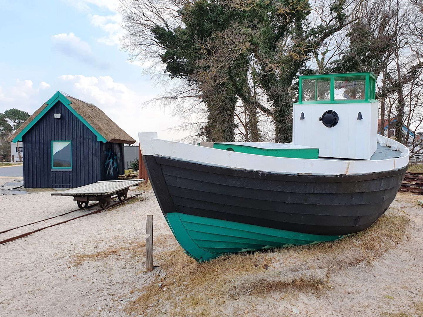 Een stilstaande boot met een groen en wit hutje, gelegen op een zandige oever met een houten constructie op de achtergrond.