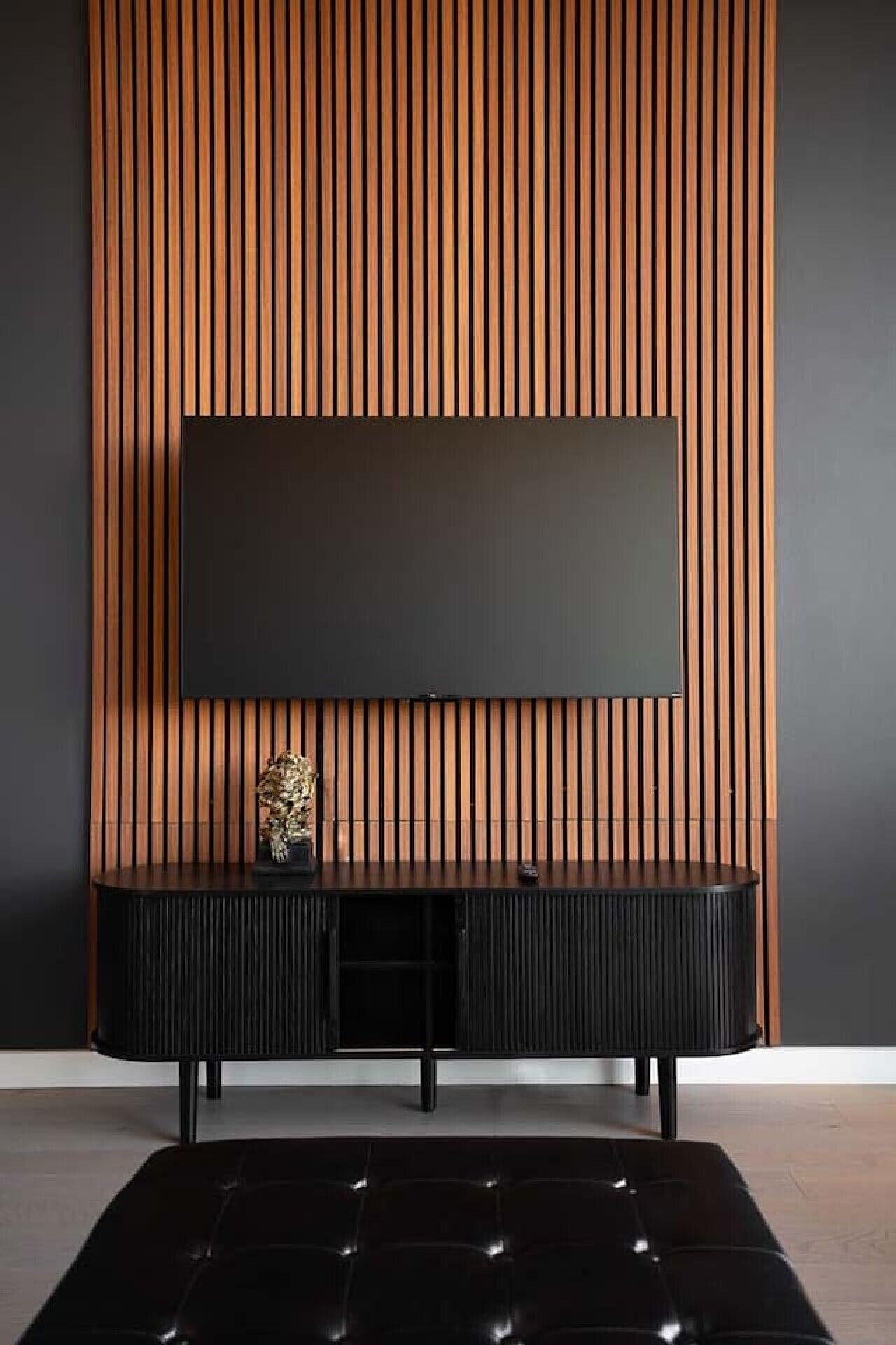 Een moderne woonkamer met een flatscreen televisie die aan een houten paneelwand is bevestigd, een zwarte entertainmentkast eronder en een zwart, gebuffeld poef op de voorgrond.