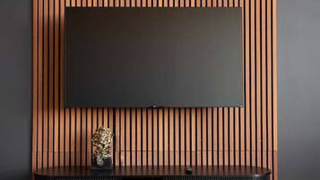 Een moderne woonkamer met een flatscreen televisie die aan een houten paneelwand is bevestigd, een zwarte entertainmentkast eronder en een zwart, gebuffeld poef op de voorgrond.