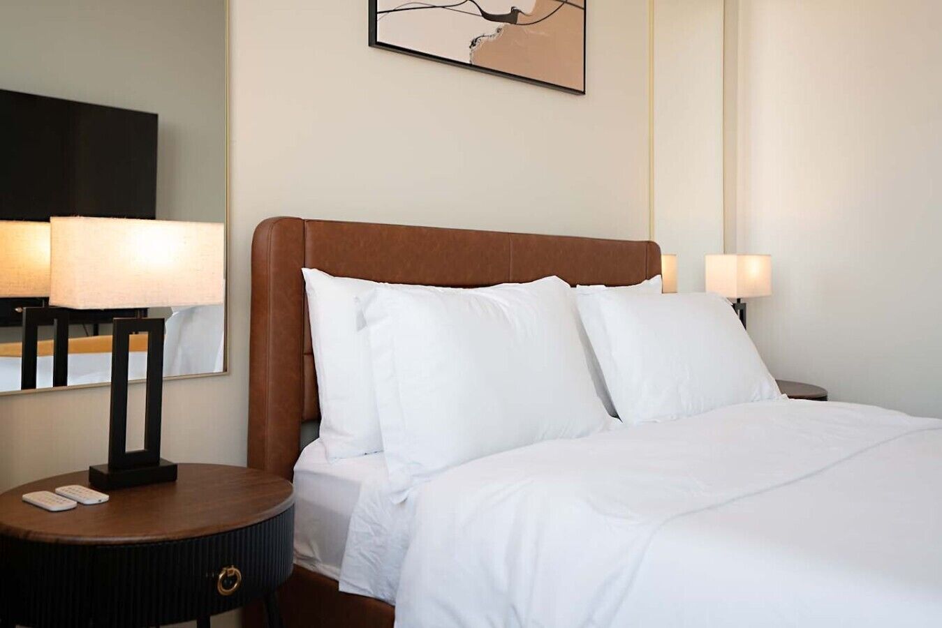 A hotel room with a bed, two bedside lamps, a nightstand with a remote control, and a wall-mounted artwork.
