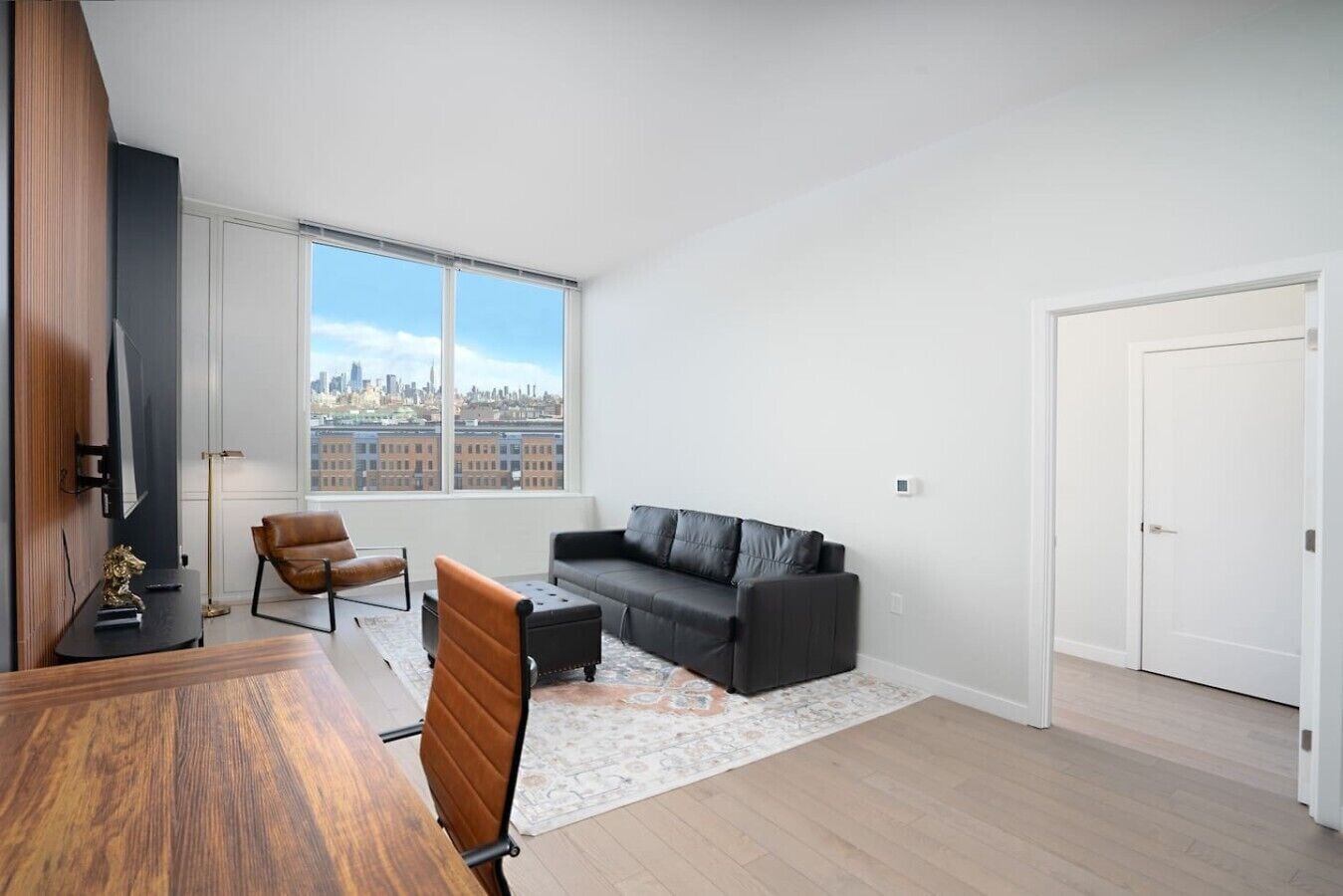 A modern living room with a large window, a black leather sofa, a brown leather chair, a wooden dining table, and a small rug.