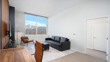 A modern living room with a large window, a black leather sofa, a brown leather chair, a wooden dining table, and a small rug.