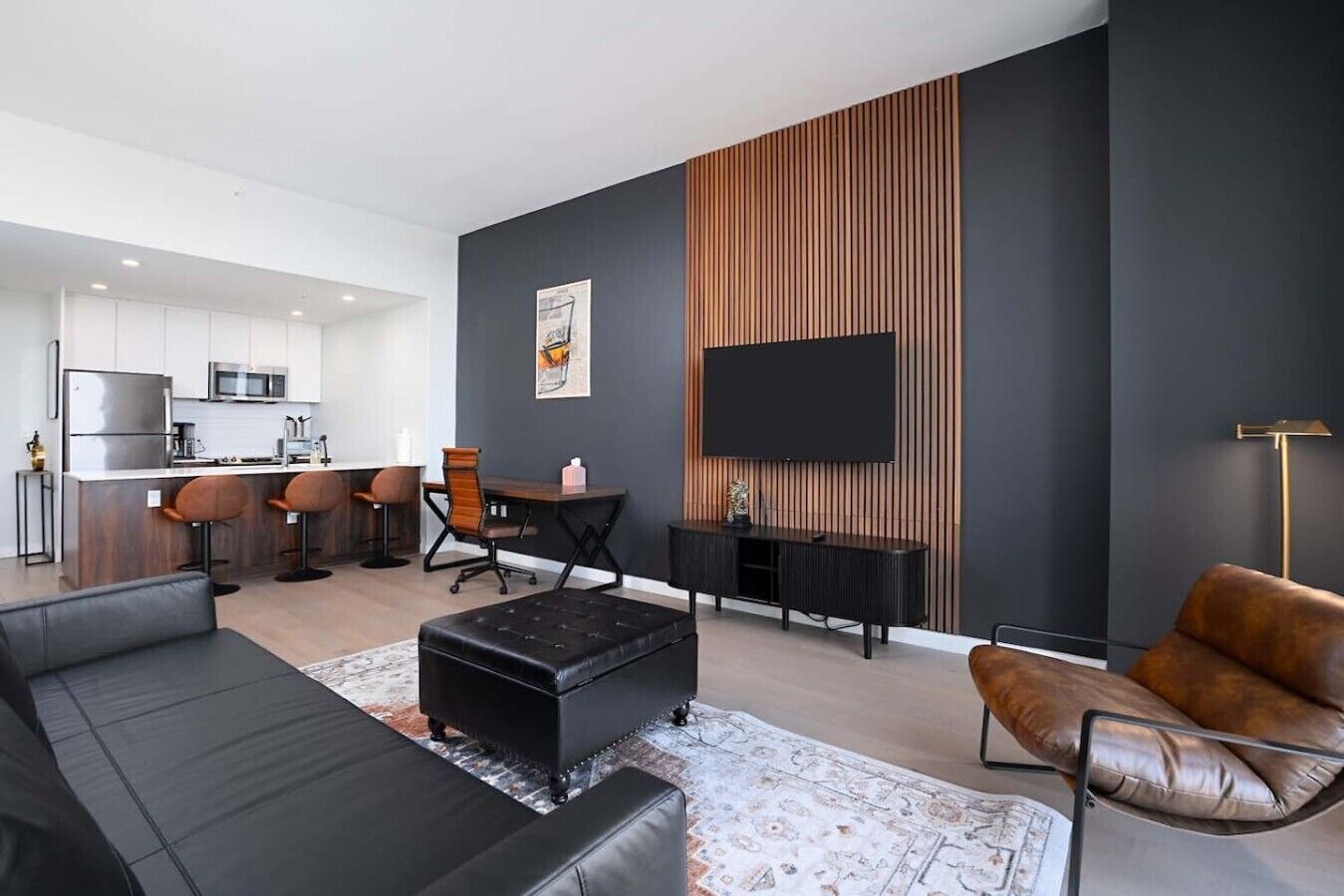 A modern living room with a black sofa, a brown leather armchair, a dining area with a wooden table and chairs, and a kitchen in the background.