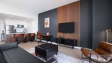 A modern living room with a black sofa, a brown leather armchair, a dining area with a wooden table and chairs, and a kitchen in the background.