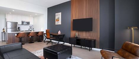 A modern living room with a black sofa, a brown leather armchair, a dining area with a wooden table and chairs, and a kitchen in the background.