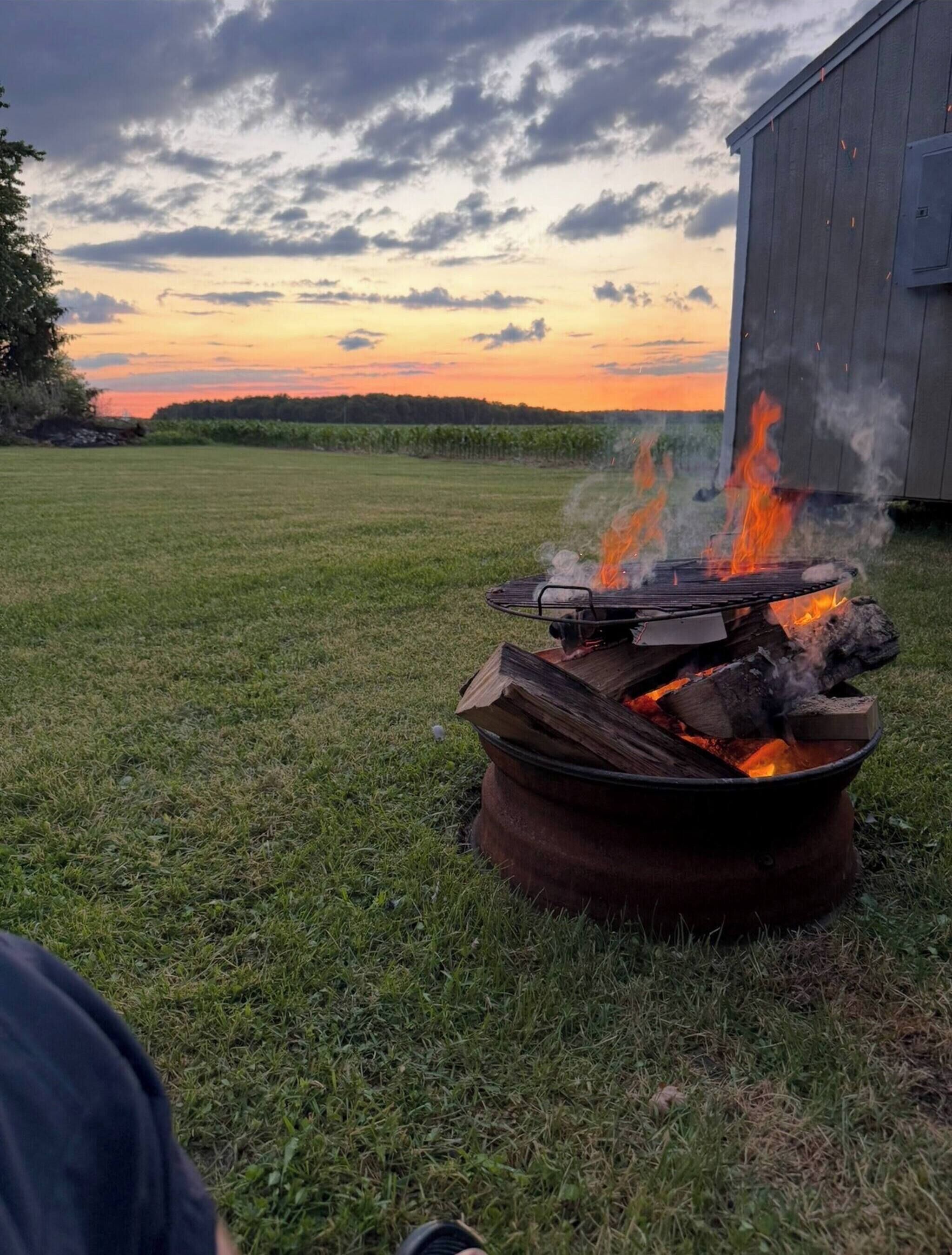 Un fogón con leña ardiendo en un campo de césped, una estructura de madera al fondo y un cielo al atardecer.