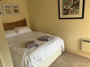 Ein ordentlich bezogenes Bett mit weißen Leinen und zwei gefalteten Handtüchern, ein hölzernes Kopfteil sowie ein gerahmtes Blumenbild an der Wand.