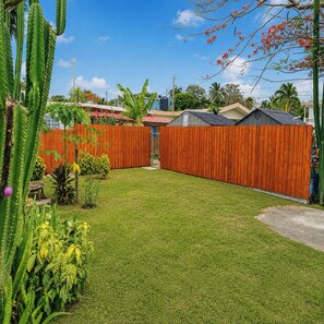 A well-maintained backyard with a wooden fence, a grassy lawn, and various plants including cacti and flowering plants. - Los Flamboyanes Cozy Suite (Aguadilla city)